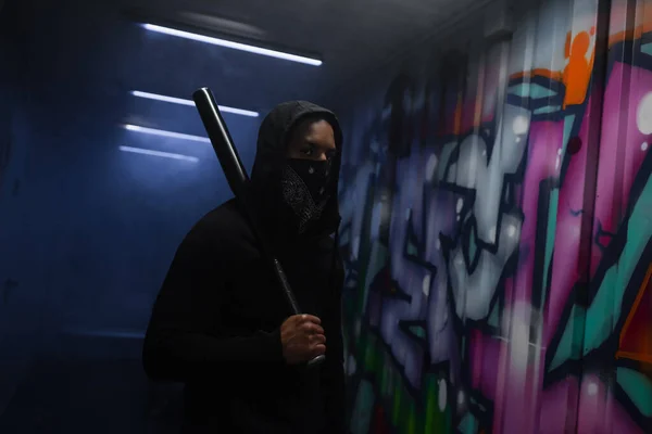 African american hooligan in mask holding baseball bat near graffiti and smoke in garage — Fotografia de Stock