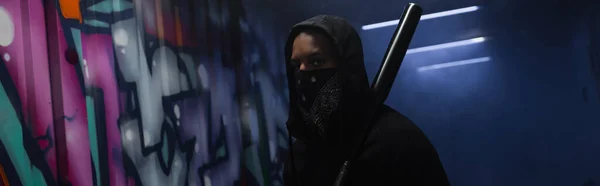 Bandido afroamericano con máscara en la cara sosteniendo bate de béisbol cerca de graffiti en el garaje con humo, pancarta - foto de stock