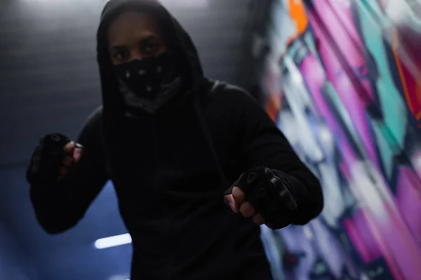 Vue en angle bas du hooligan afro-américain flou dans les gants et le masque debout dans la pose de combat dans le garage — Stock Photo