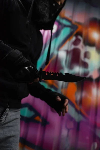 Vista recortada del cuchillo de mano de ladrón afroamericano cerca de graffiti borroso en la pared al aire libre - foto de stock