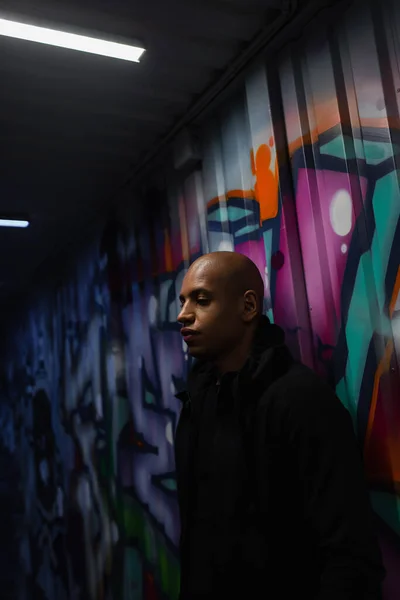 African american hooligan standing near graffiti in garage - foto de stock