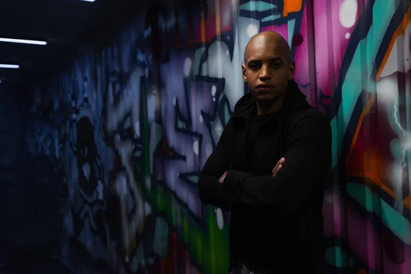 African american hooligan looking at camera near graffiti in garage - foto de stock