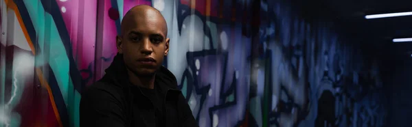 African american hooligan looking at camera near graffiti on wall in garage, banner — Photo de stock