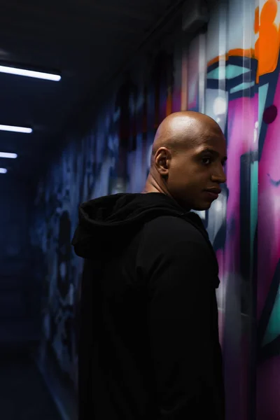 African american hooligan in hoodie looking away near graffiti on wall in garage — Stock Photo