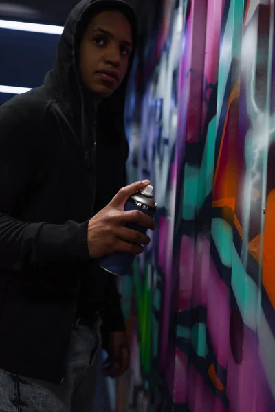 Vândalo afro-americano desfocado na pintura do capuz graffiti e olhando para longe na garagem — Fotografia de Stock