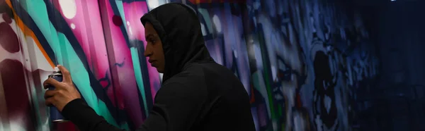 Side view of african american vandal painting graffiti on wall on urban street at night, banner — Photo de stock