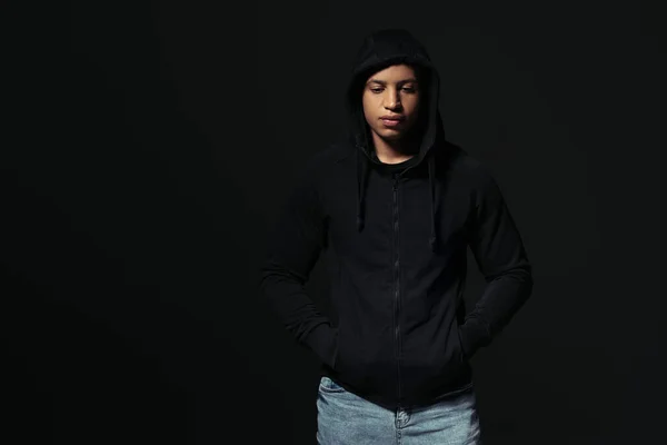 African american hooligan holding hands in pockets of hoodie isolated on black — Fotografia de Stock