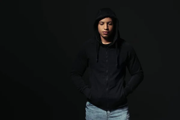 African american hooligan holding hands in pockets of hoodie isolated on black — Foto stock