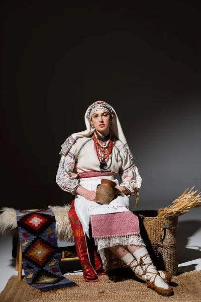 Full length of young ukrainian woman in traditional clothing with ornament holding clay pot while sitting on bench on black — Photo de stock