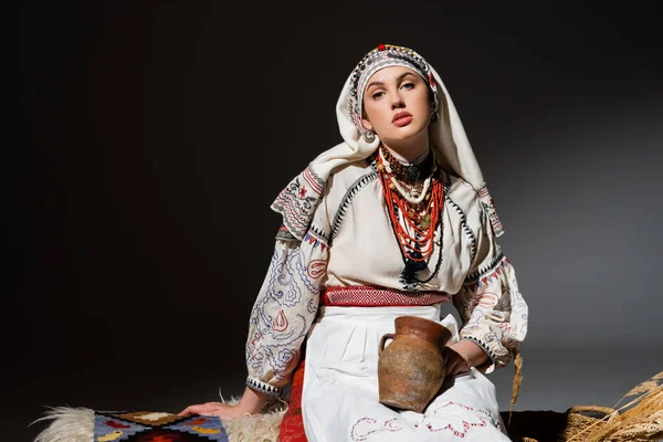 Юна українка в традиційному одязі з орнаментом, що тримає глиняний горщик і сидить на лавці на чорному. — стокове фото