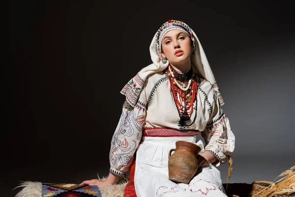 Young ukrainian woman in traditional clothing with ornament holding clay pot and sitting on bench on black — Stock Photo