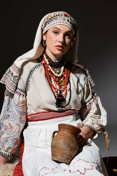 Pretty ukrainian woman in traditional clothing with ornament holding clay pot on black — Photo de stock