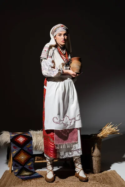 Full length of pretty  ukrainian woman in traditional clothing with ornament holding clay pot while standing on black — Stockfoto