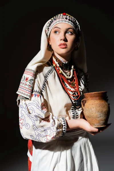 Portrait of pretty ukrainian woman in traditional clothing with ornament holding clay pot on black — Stockfoto