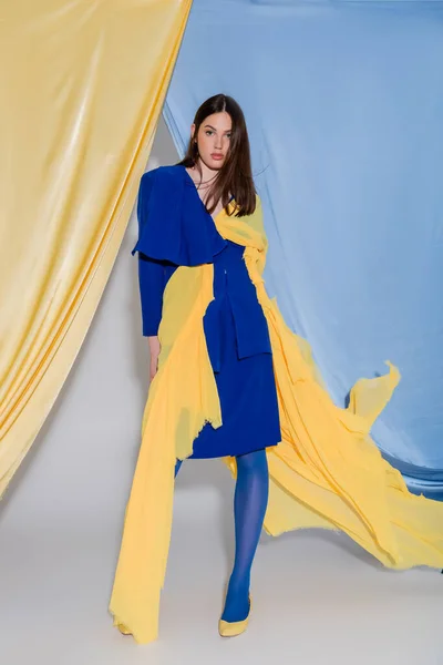 Pleine longueur de jeune femme ukrainienne en robe de bloc de couleur posant près de rideaux bleus et jaunes — Photo de stock