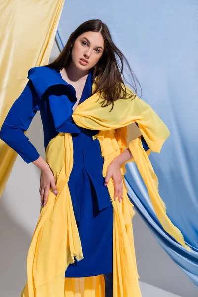 Young ukrainian woman in color block dress posing with hands on hips near blue and yellow curtains - foto de stock