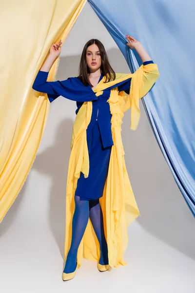 Полная длина красивой украинской женщины в цветном блоке платье позирует рядом с синими и желтыми занавесками — стоковое фото