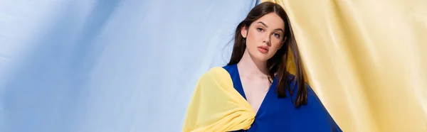 Young ukrainian woman in color block clothes looking at camera near blue and yellow curtains, banner - foto de stock