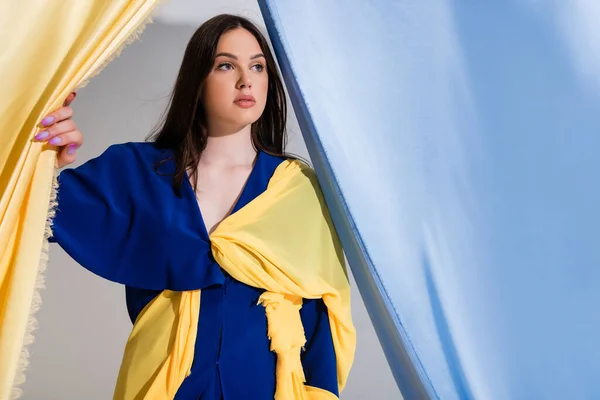 Nachdenkliche junge Ukrainerin im Farbblockkleid posiert in der Nähe blauer und gelber Vorhänge — Stockfoto