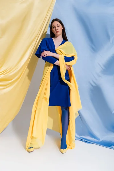 Pleine longueur de confiance jeune femme ukrainienne en robe de bloc de couleur posant près de rideaux bleus et jaunes — Photo de stock