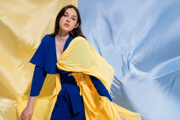 Brunette ukrainian woman in color block clothing posing near blue and yellow fabric — Foto stock