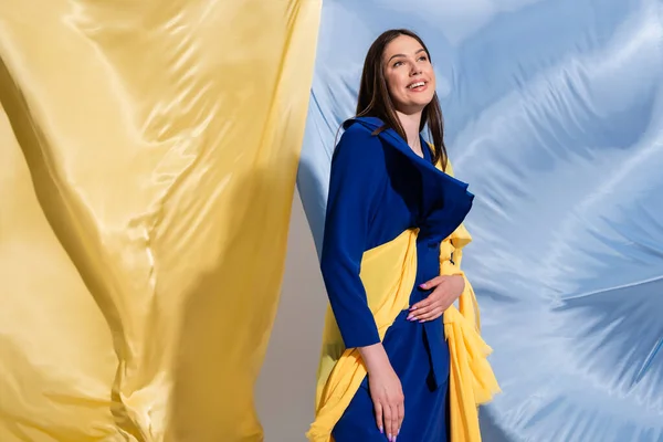 Joyful young ukrainian woman in color block clothing posing near blue and yellow fabric — Foto stock