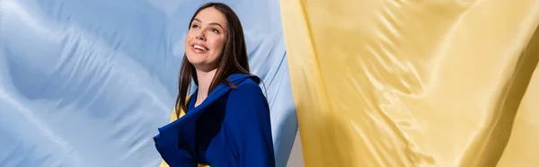 Cheerful young ukrainian woman in color block clothing posing near blue and yellow flag, banner — Stock Photo