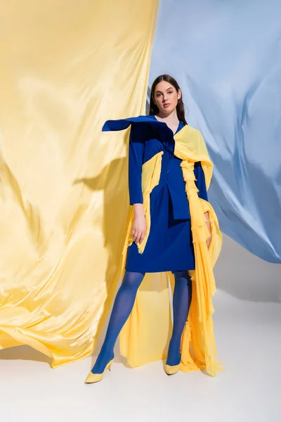 Full length of young ukrainian woman in fashionable color block clothing posing near blue and yellow fabric — Stock Photo