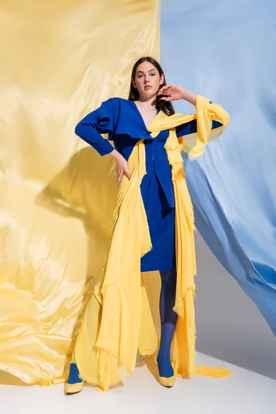 Volle Länge der ukrainischen Frau in modischen Color Block Kleidung posiert mit der Hand auf der Hüfte in der Nähe von blauen und gelben Stoff — Stockfoto