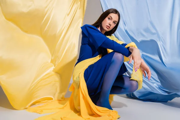 Mujer ucraniana joven en ropa de bloque de color elegante sentado cerca de tela azul y amarillo - foto de stock