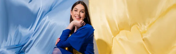 Positive ukrainian woman in stylish color block clothing posing near blue and yellow flag, banner — Photo de stock