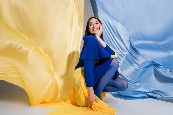 Cheerful ukrainian woman in stylish color block clothing sitting near blue and yellow fabric — Foto stock