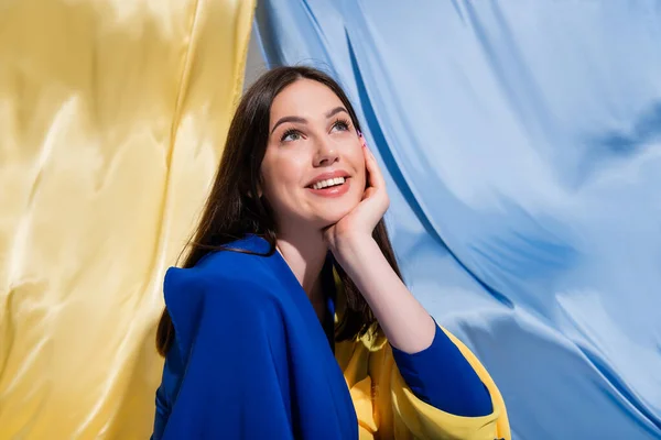 Dreamy ukrainian woman in stylish color block clothing smiling near blue and yellow flag — Stock Photo