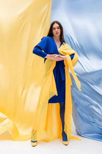 Volle Länge der ukrainischen junge Frau in stilvoller Color Block Kleidung posiert in der Nähe von blauen und gelben Stoff — Stockfoto