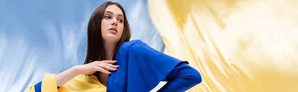 Ukrainian young woman in stylish color block clothing posing near blue and yellow fabric, banner — Photo de stock