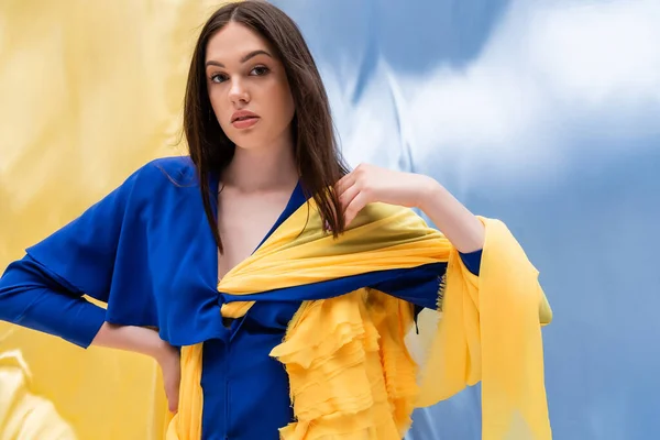 Brünette junge Frau in stylischer Color-Block-Kleidung posiert mit der Hand auf der Hüfte nahe der ukrainischen Flagge — Stockfoto