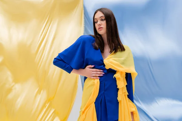 Pretty ukrainian young woman in stylish color block clothing standing with hand on hip near blue and yellow fabric — Foto stock