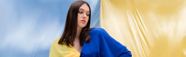 Гарна українська молода жінка в стильному одязі позує біля синьо-жовтого прапора, банер — стокове фото
