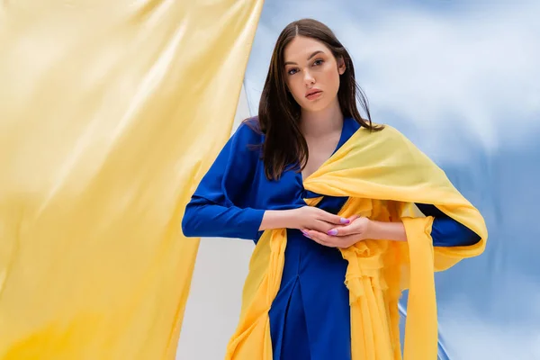 Патриотическая украинская молодая женщина в стильной одежде позирует рядом с сине-желтой тканью — стоковое фото