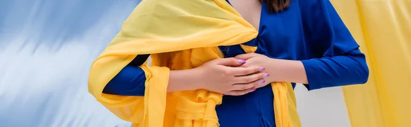Vue recadrée de la jeune femme ukrainienne patriotique dans des vêtements élégants posant près de tissu bleu et jaune, bannière — Photo de stock