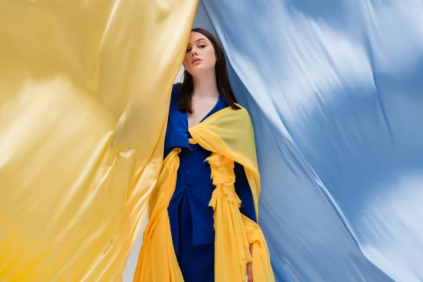 Patriotic ukrainian young woman in fashionable clothing posing near blue and yellow fabric — Photo de stock