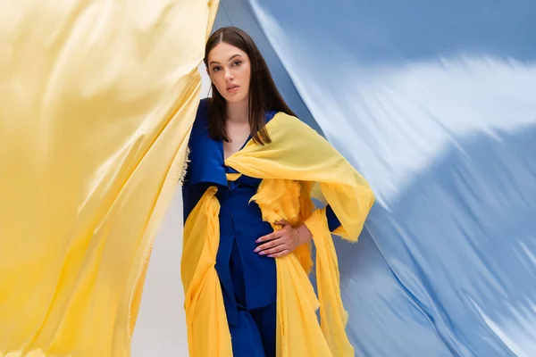 Patriotic ukrainian young woman in trendy clothing posing with hand on hip near blue and yellow fabric — Foto stock