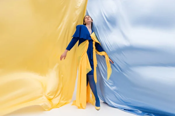 Comprimento total da mulher ucraniana patriótica na roupa na moda posando perto de tecido azul e amarelo — Fotografia de Stock