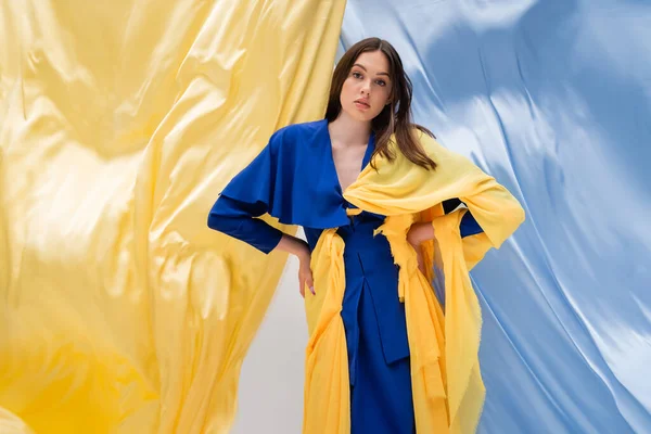 Patriotic ukrainian model in trendy outfit posing with hands on hips near blue and yellow drapery — Stockfoto