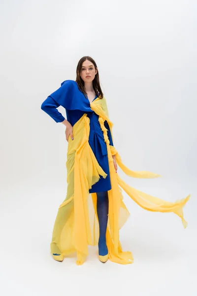 Pleine longueur de femme ukrainienne élégante en tenue bleue et jaune posant avec la main sur la hanche sur le gris — Photo de stock
