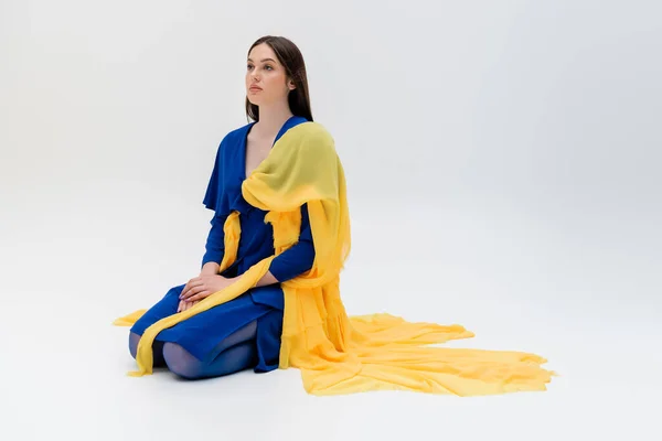 Pleine longueur de femme ukrainienne patriotique en tenue bleue et jaune assis sur gris — Photo de stock