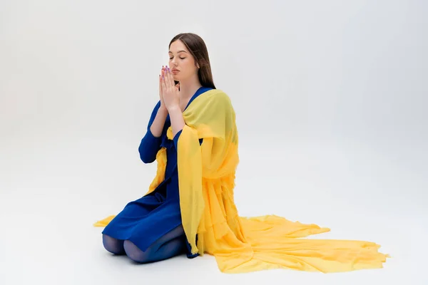 Full length of stylish ukrainian woman in blue and yellow outfit praying on grey — Photo de stock