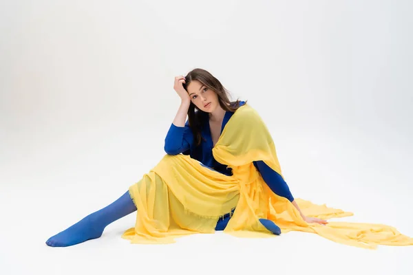 Patriotic ukrainian young woman in blue and yellow clothes sitting while posing on grey — Stockfoto