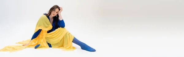 Patriotic ukrainian young woman in blue and yellow clothes sitting while posing on grey, banner — Stockfoto