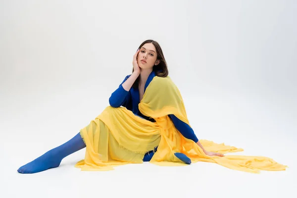 Patriotic ukrainian young woman in blue and yellow outfit with tights sitting while posing on grey — Photo de stock